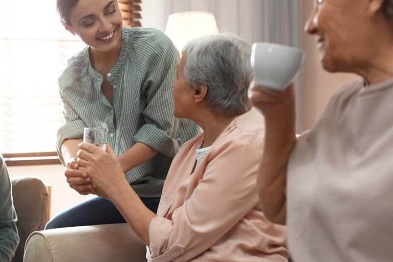 Young woman taking care of elderly people in living room Caregiver helps senior living residents drink from cup
