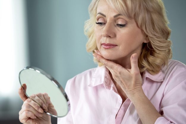 Attractive aged woman looking at her face in mirror, plastic surgery effect