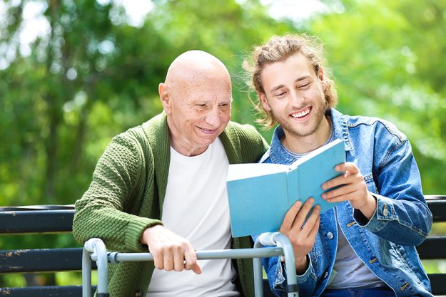 Young man reading book to his elderly father in park Young man reading book to his elderly father in park