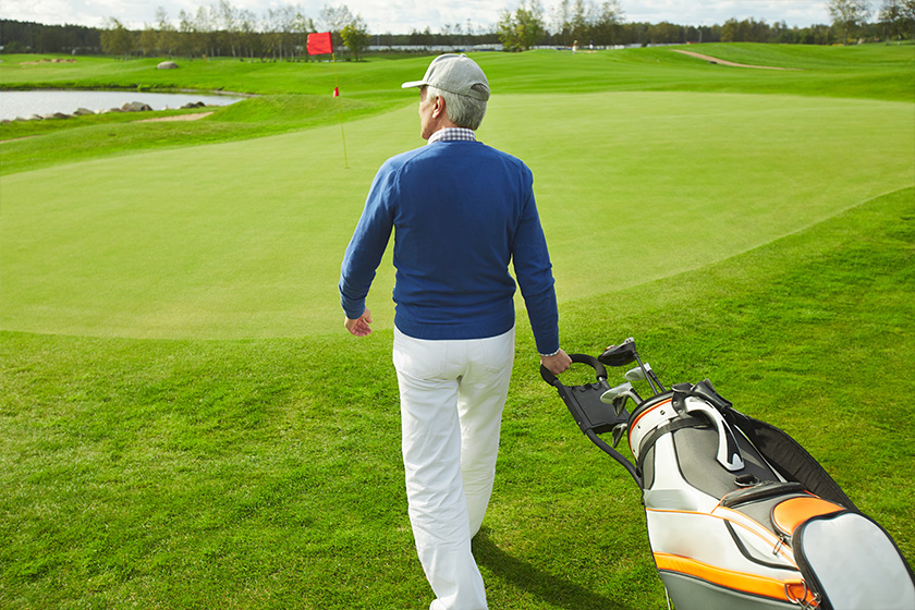 senior man walking down green field and carrying bag with bunch of golf clubs