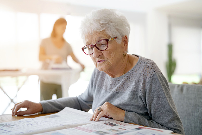 Portrait of old woman reading newspaper while homehelp irons