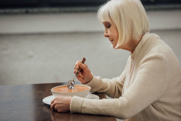 sad senior woman sitting at table and eating cream soup at home
