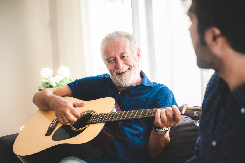 happy adult son and senior father playing guitar on sofa at home relaxing and having happy time with family Are There Any Requirements For Staying In Senior Living Communities In Griffith, IN?