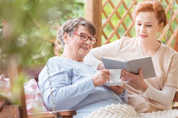 Happy elderly woman drinking tea while nurse reading book in the garden Happy elderly woman drinking tea while nurse reading book in the garden
