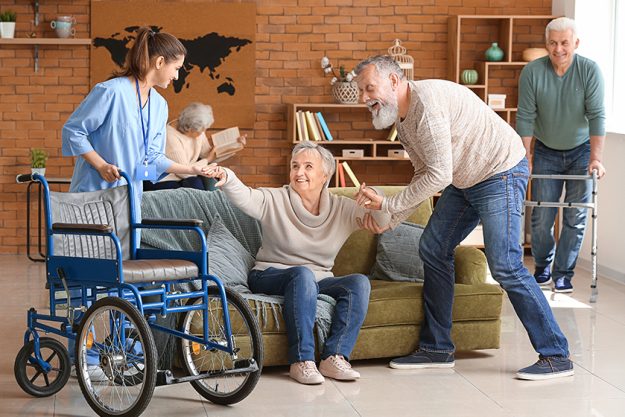 Caregiver and elderly man helping senior woman to sit in wheelchair