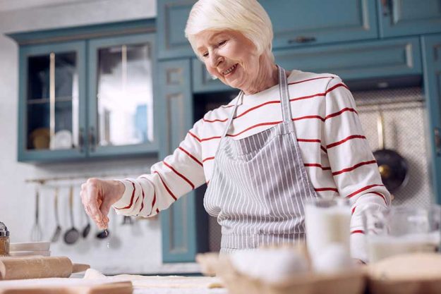 Cheerful senior woman making dough in kitchen