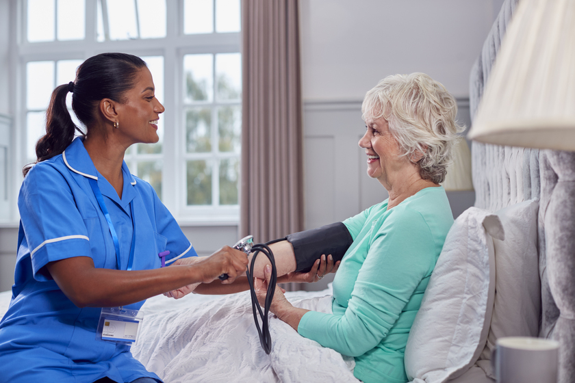 Senior Woman At Home In Bed Having Blood Pressure Taken By Female Care Worker In Uniform Signs Your Aging Parents Are Struggling With Daily Routines And Need Senior Personal Care Services In Fogelsville, PA
