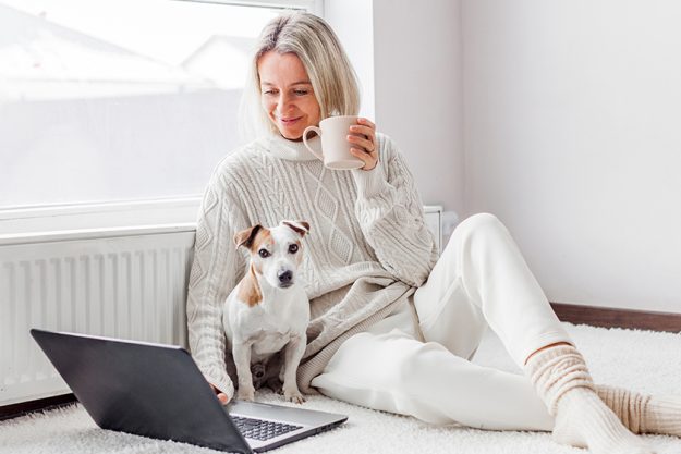 Relaxed middle-aged woman is working on a laptop at her home. Adult female is sitting on the floor near the radiator in a white knitted sweater with her dog and watching movie