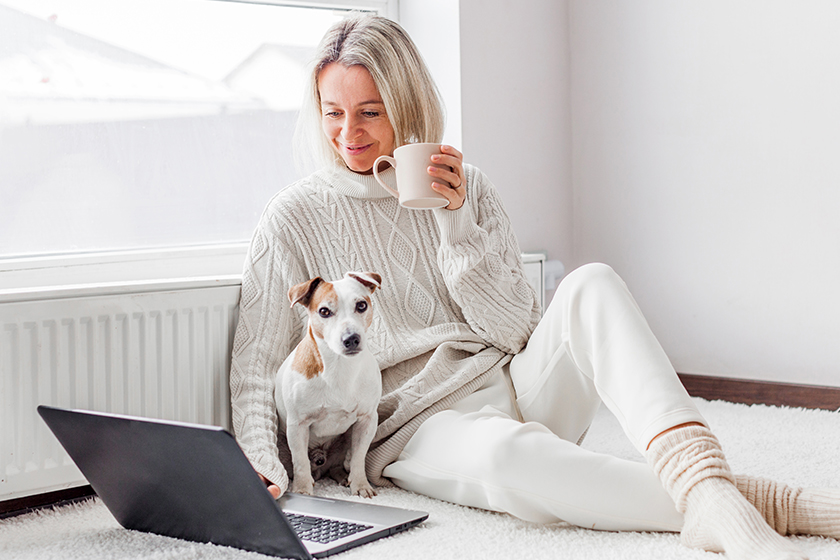 Relaxed middle-aged woman is working on a laptop at her home. Adult female is sitting on the floor near the radiator in a white knitted sweater with her dog and watching movie