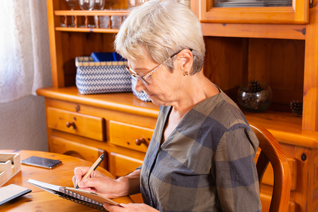 Senior mature Caucasian woman making notes while sitting near the table Senior mature Caucasian woman making notes while sitting near the table