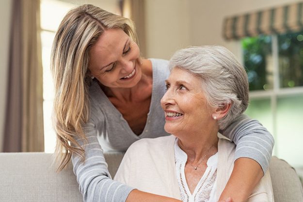 Portrait of old mother and mature daughter hugging at home. Happy senior mother and adult daughter embracing with love on sofa. Cheerful woman hugging from behind older mom and looking at each other.