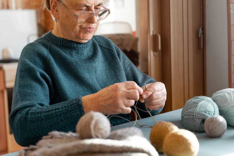 An elderly man with poor eyesight is engaged in knitting while sitting at home in the kitchen. There are balls of blue, brown and gray threads on the table. Focus on the face. An elderly man with poor eyesight is engaged in knitting while sitting at home in the kitchen. There are balls of blue, brown and gray threads on the table. Focus on the face.