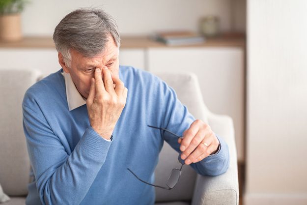Senior Man Massaging Nosebridge Sitting On Sofa