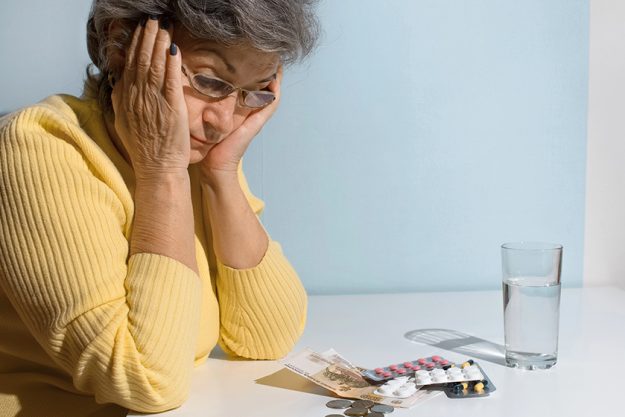 Elderly woman with glasses looking at money and pills on the table. Concept of depression, drug price, treatment cost Elderly woman with glasses looking at money and pills on the table. Concept of depression, drug price, treatment cost
