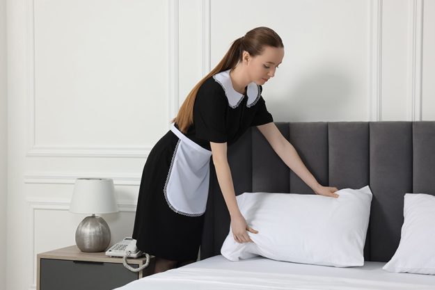Young maid making bed in hotel room