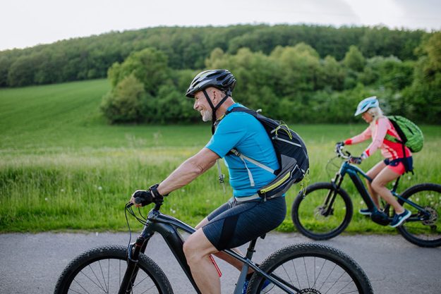 An active senior couple riding electric bicycles on path at summer park, healthy lifestyle concept. Side view.