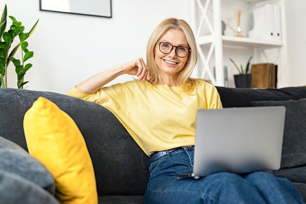 Caucasian senior woman spending home leisure with laptop. Calm woman sitting at cozy sofa and looking at camera. Web browsing, watching TV series
