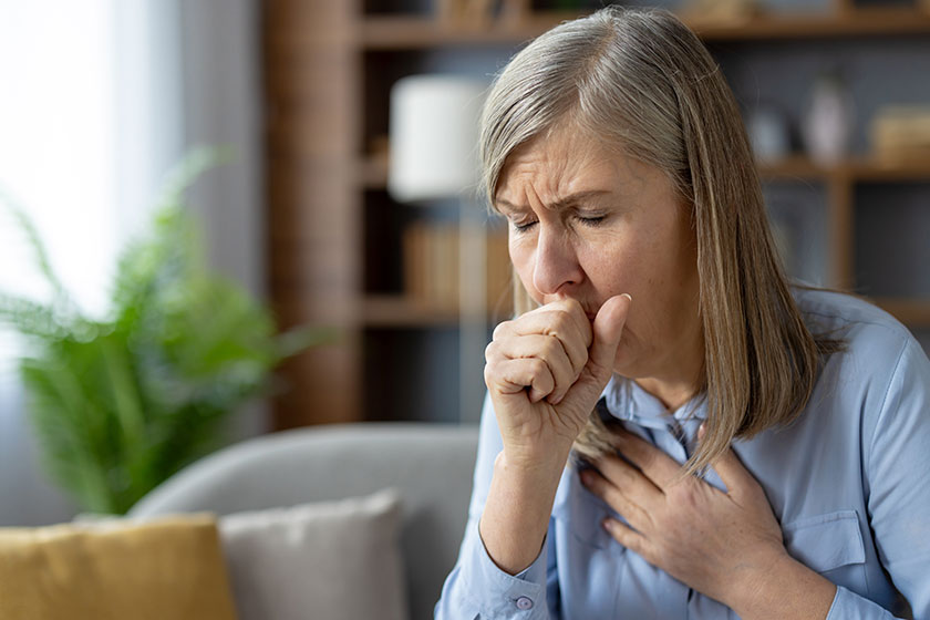 An older woman looks distressed as she coughs, clutching her chest in discomfort. She is sitting in a well-furnished living room, expressing signs of illness.