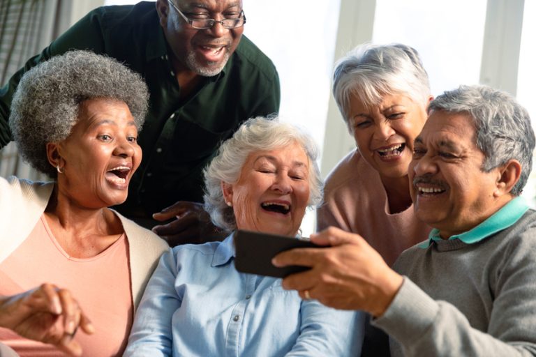 Five happy diverse senior friends sitting on sofa and looking at smartphone. retirement lifestyle relaxing at home with technology.