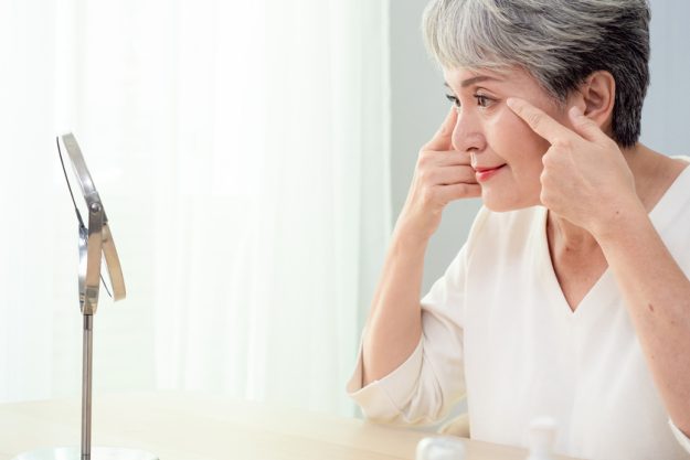 Happy 50s middle aged asian woman touching face skin looking in mirror reflection. Smiling mature old lady pampering, healthy moisturized skin care, aging beauty, skincare treatment cosmetics concept. Happy 50s middle aged asian woman touching face skin looking in mirror reflection. Smiling mature old lady pampering, healthy moisturized skin care, aging beauty, skincare treatment cosmetics concept.