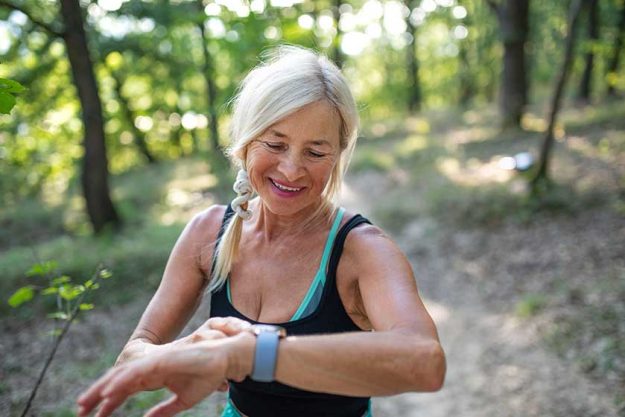 Portrait-of-active-senior-woman-runner-standing-outdoors-in-forest-setting-smartwatch