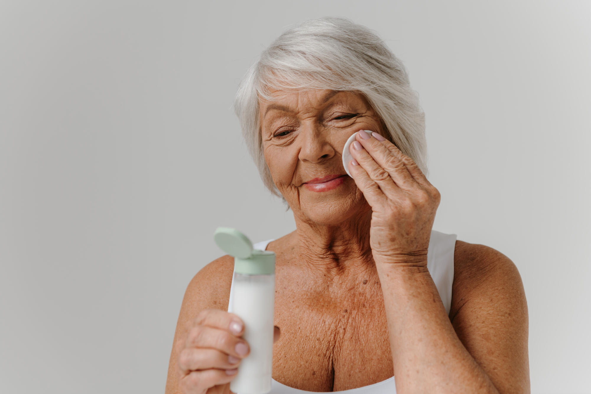 Confident senior woman cleaning face with cotton sponge against grey background