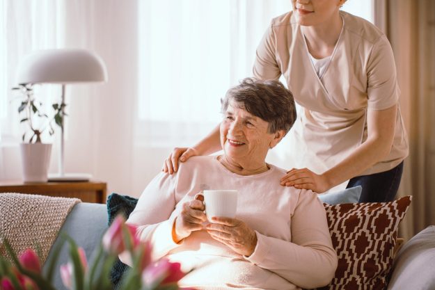 Smiling,Senior,Woman,Drinking,Tea,With,Her,Caregiver,Standing,Behind
