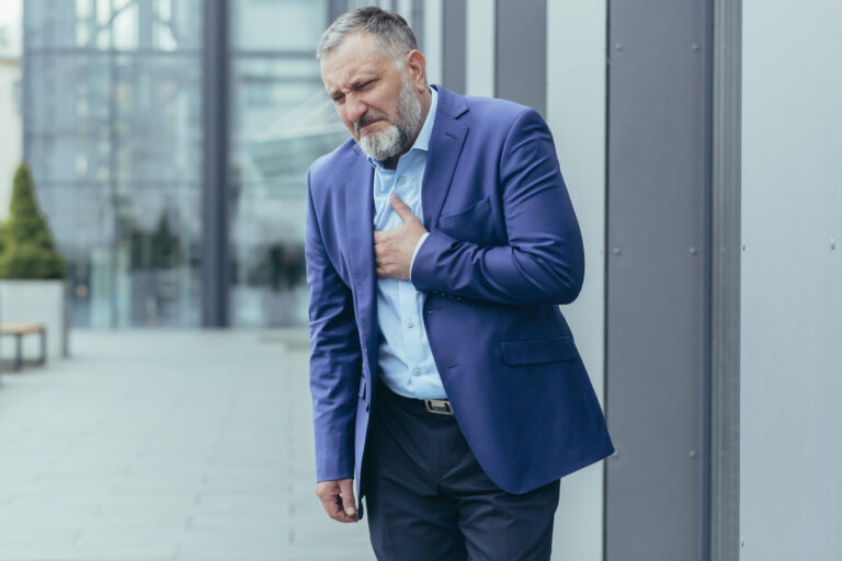 Pre-infarction condition. A senior man in a suit stands on the street near an office center holding his heart. He feels a sharp pain in his chest. He needs urgent medical help.