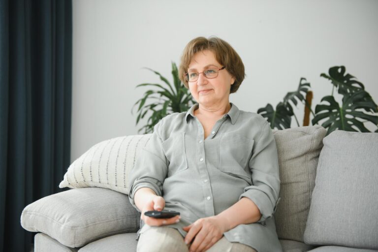 people, television and entertainment concept – senior woman in eyeglasses with remote control watching tv at home